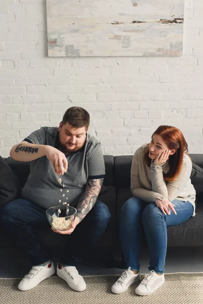 Fidanzata guardando come fidanzato in sovrappeso giocare con popcorn a casa — Foto stock