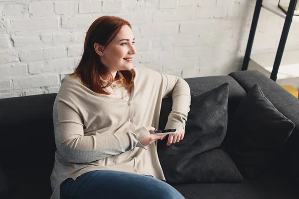 Lächelndes schönes Mädchen, das Smartphone in der Hand hält und zu Hause wegschaut — Stockfoto