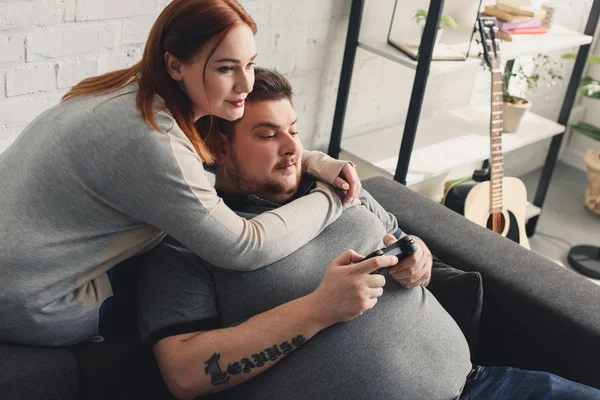 Taille plus petite amie étreignant petit ami pendant qu'il joue à un jeu vidéo à la maison — Stock Photo