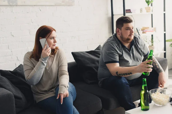 Sobrepeso novia hablando por teléfono inteligente y mirando novio con cerveza en casa - foto de stock