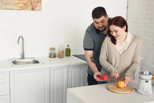 Boyfriend presenting gift to girlfriend while she cutting orange at kitchen — Stock Photo