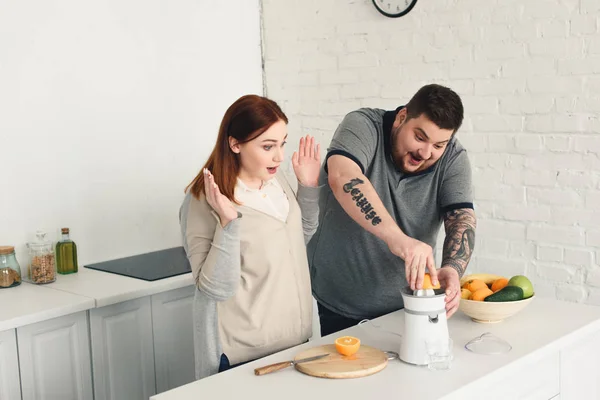 Surprised size plus girlfriend looking how boyfriend preparing orange juice at home — Stock Photo