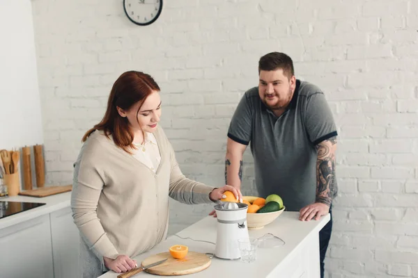 Novio con sobrepeso y novia preparando jugo de naranja en la cocina - foto de stock