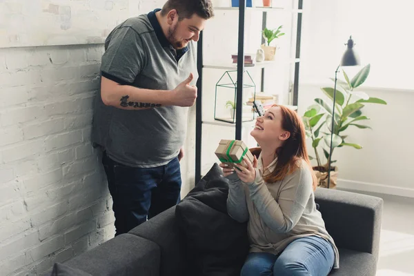 Freund zeigt Daumen hoch und lächelnde Freundin hält Geschenkbox zu Hause — Stockfoto