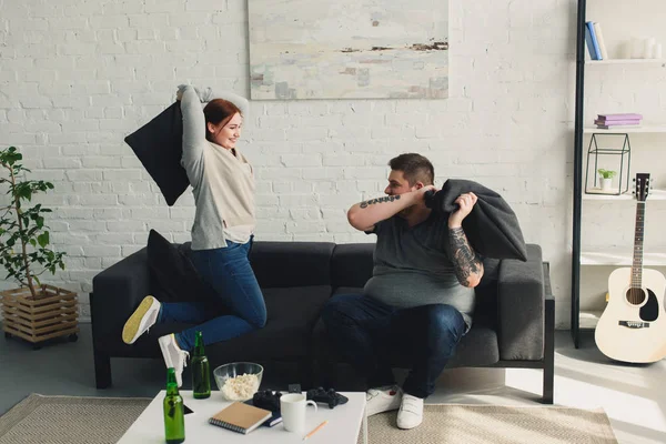 Novio y novia luchando con almohadas en la sala de estar - foto de stock