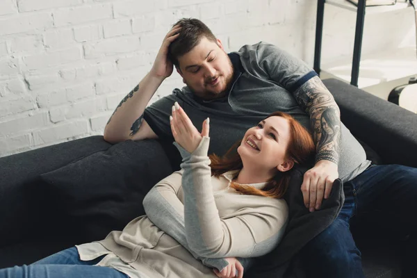 Smiling size plus boyfriend and girlfriend talking and lying on sofa at home — Stock Photo