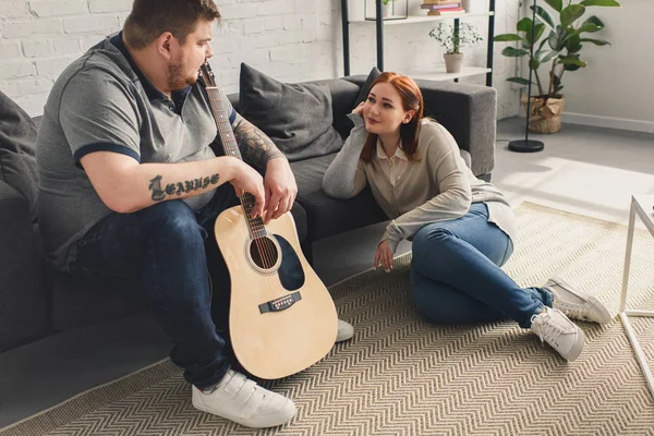 Tamaño más novio sosteniendo la guitarra acústica y mirando a la novia en casa - foto de stock