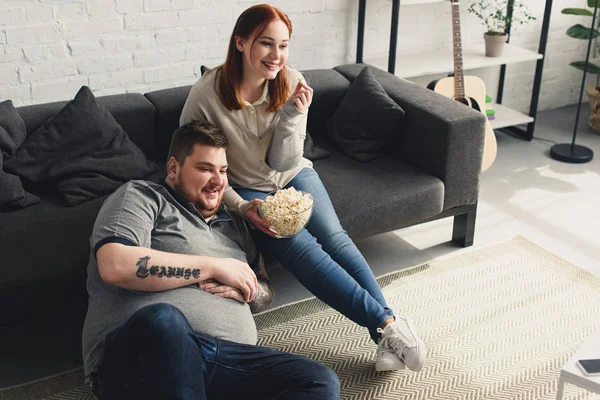 Smiling overweight boyfriend and girlfriend eating popcorn and watching tv at home — Stock Photo