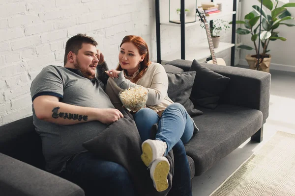Tamaño más novia alimentación novio con palomitas de maíz en el sofá en casa - foto de stock