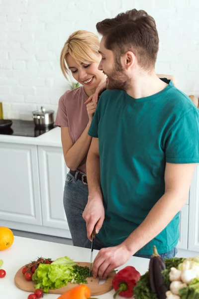 Veganer Freund schneidet Gemüse und Freundin umarmt ihn in der Küche — Stockfoto