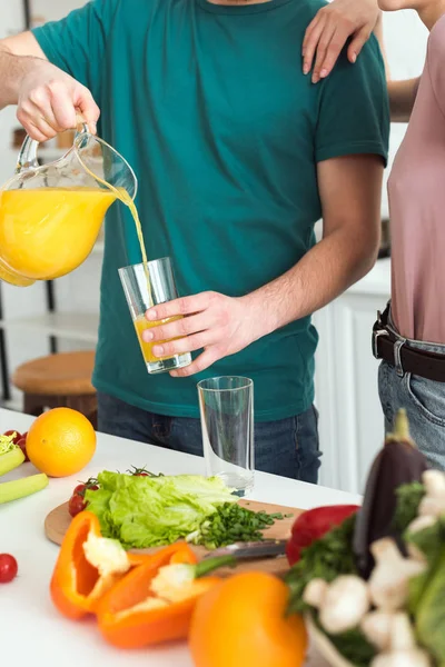 Imagen recortada de novio vegano vertiendo jugo fresco en el vidrio en la cocina - foto de stock