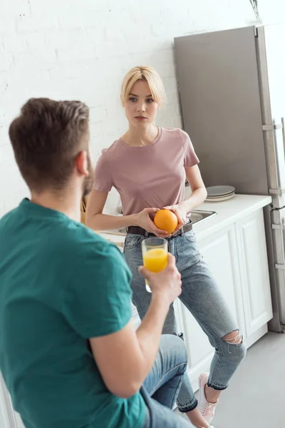 Pareja de veganos hablando en la cocina y sosteniendo jugo de naranja y naranja - foto de stock