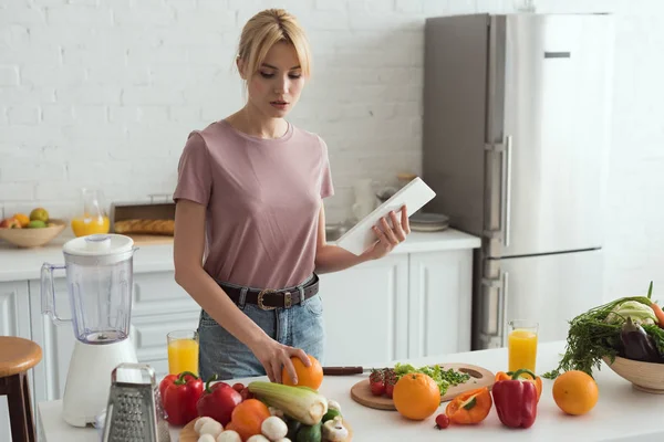 Attractive vegan girl cooking and holding tablet with recipe in kitchen — Stock Photo
