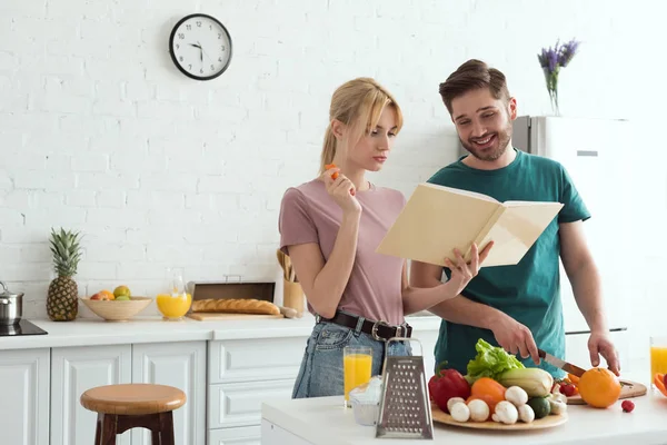 Couple of vegans preparing salad with recipe book at kitchen — Stock Photo