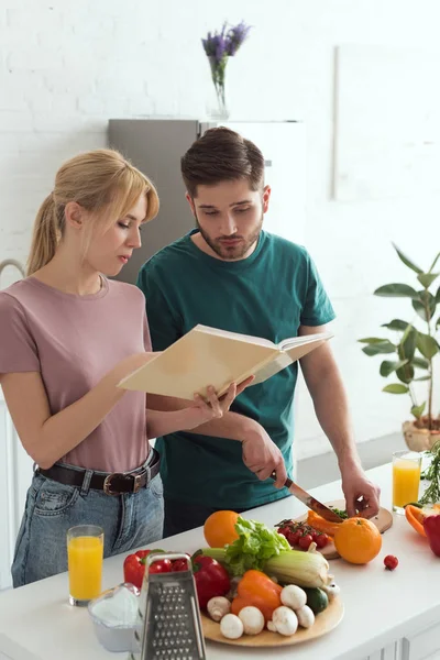 Pareja de veganos preparando comida con libro de cocina en la cocina - foto de stock