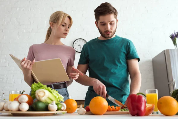 Casal de vegans preparando alimentos com livro de receitas na cozinha — Fotografia de Stock