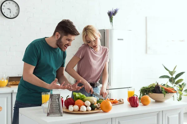 Novio haciendo muecas mientras su novia cocina y hablando por teléfono inteligente en la cocina - foto de stock