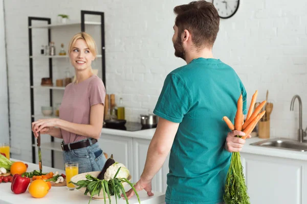 Vegan boyfriend hiding bouquet of carrots from girlfriend at kitchen — Stock Photo