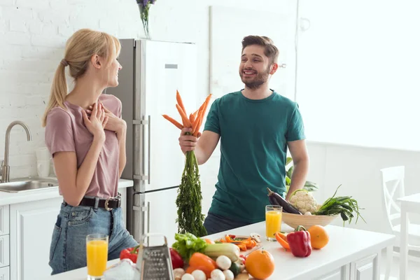 Vegan boyfriend presenting bouquet of carrots to girlfriend at kitchen — Stock Photo