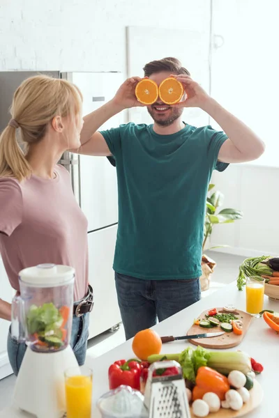 Fidanzato divertirsi e coprire gli occhi con pezzi di arancia in cucina, concetto vegan — Foto stock