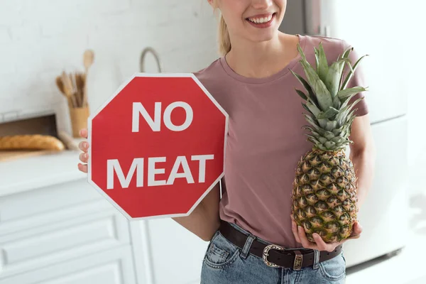 Vue partielle de la femme souriante ne tenant aucun signe de viande et d'ananas frais dans les mains dans la cuisine, concept de mode de vie végétalien — Photo de stock