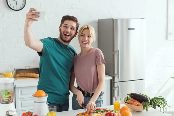 Улыбающаяся веганская пара делает селфи, готовясь вместе на кухне дома — стоковое фото