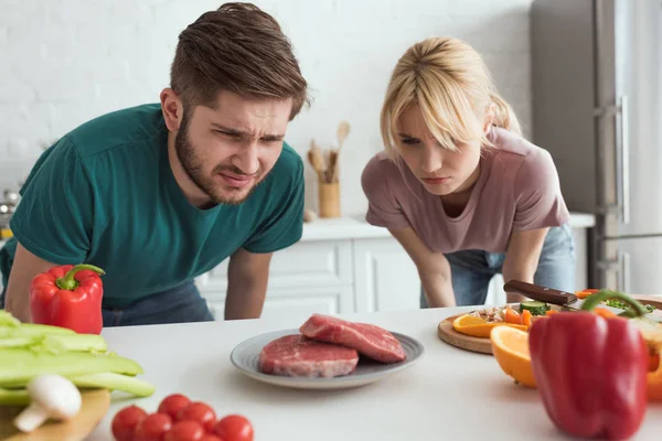 Disgusted vegan couple looking at raw meat on plate in kitchen at home — Stock Photo
