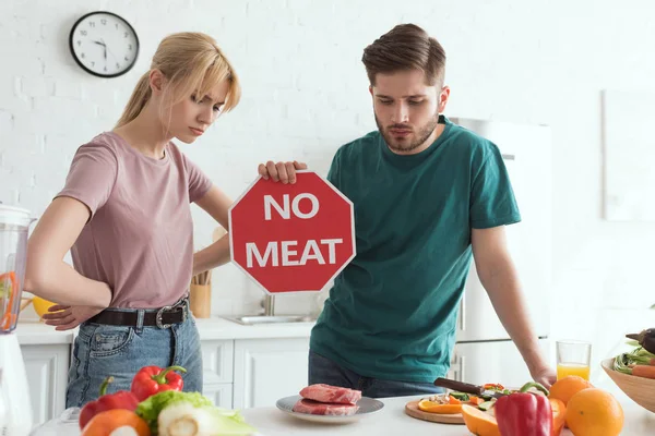 Vegan couple with no meat sign at table with raw meat and vegetables in kitchen at home — Stock Photo