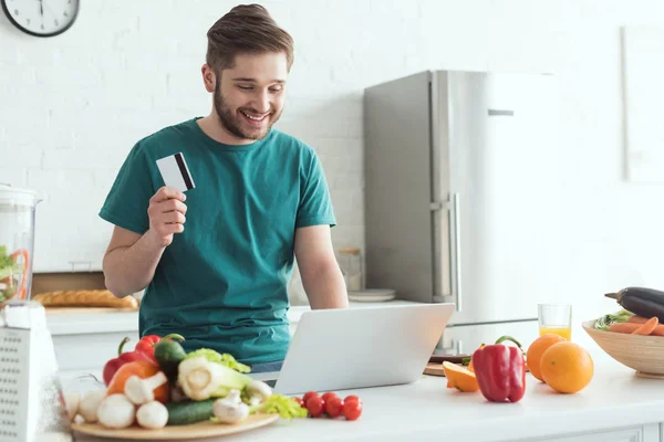 Uomo sorridente con carta di credito e laptop acquistare beni online in cucina a casa — Foto stock