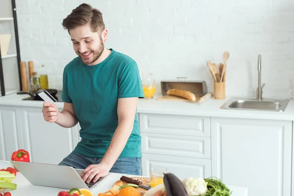 Homme souriant avec carte de crédit et ordinateur portable acheter des marchandises en ligne dans la cuisine à la maison — Photo de stock