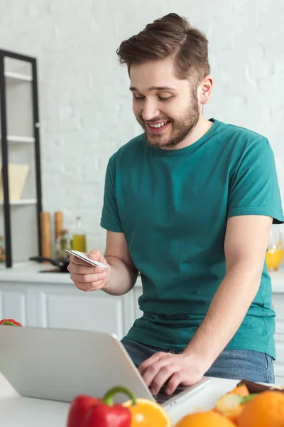 Улыбающийся мужчина с кредитной картой и ноутбуком, покупающий товары онлайн на кухне дома — стоковое фото