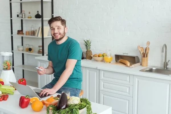 Hombre sonriente con tarjeta de crédito sentado en la mesa con el ordenador portátil en la cocina en casa - foto de stock