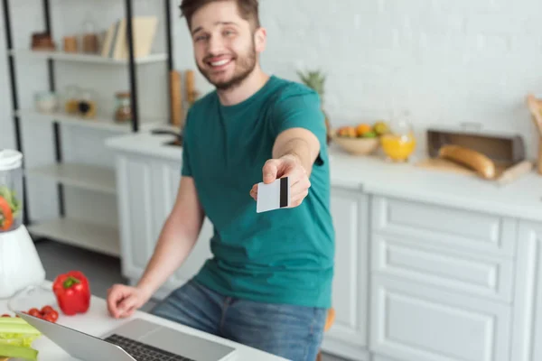 Homme souriant montrant carte de crédit à table avec ordinateur portable dans la cuisine à la maison — Photo de stock