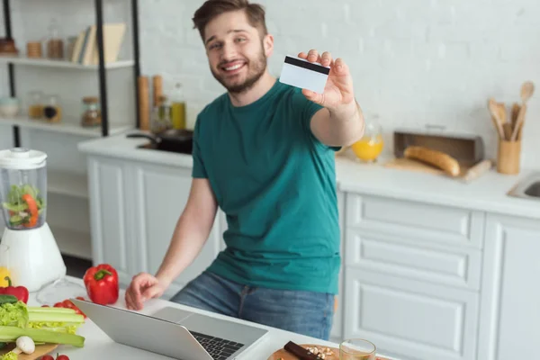 Smiling man showing credit card at table with laptop in kitchen at home — Stock Photo