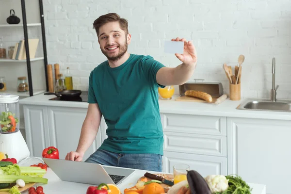 Uomo sorridente che mostra carta di credito a tavola con computer portatile in cucina a casa — Foto stock