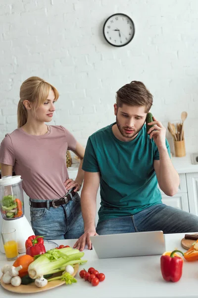 Homem fingindo falar sobre pepino à mesa com laptop e namorada perto na cozinha em casa, conceito de estilo de vida vegan — Fotografia de Stock