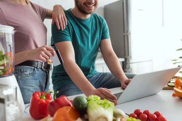 Vue partielle du couple végétalien avec ordinateur portable shopping en ligne dans la cuisine à la maison — Photo de stock