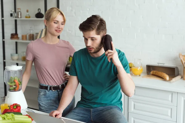 Man pretending talking on eggplant at table with laptop and girlfriend near by in kitchen at home, vegan lifestyle concept — Stock Photo