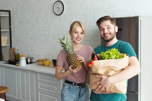 Улыбающаяся веганская пара с бумажным пакетом, полным свежих овощей на кухне дома — стоковое фото