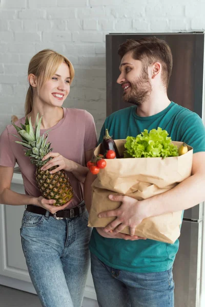 Sonriente pareja vegana con bolsa de papel llena de verduras frescas en la cocina en casa - foto de stock