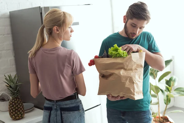 Pareja vegana con bolsa de papel llena de verduras frescas en la cocina en casa - foto de stock