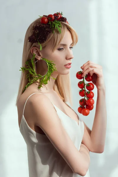 Vue latérale de la jeune femme aux tomates cerises à la main, couronne de légumes frais et boucle d'oreille en roquette fraîche, concept de mode de vie végétalien — Photo de stock