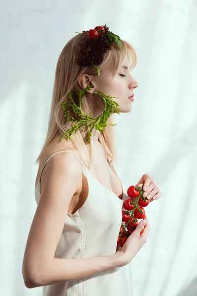 Vista lateral da jovem com tomates cereja nas mãos, grinalda feita de legumes frescos e brinco feito de arugula fresca, conceito de estilo de vida vegan — Fotografia de Stock