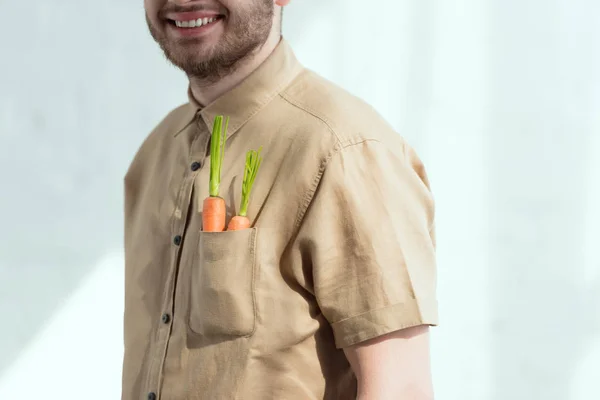 Обрезанный снимок улыбающегося бородатого человека со свежей морковью в кармане, концепция веганского образа жизни — стоковое фото