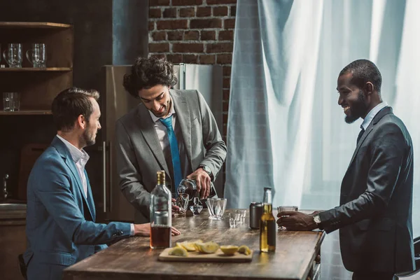 Tres jóvenes sonrientes hombres multiétnicos en trajes que beben bebidas alcohólicas juntos - foto de stock