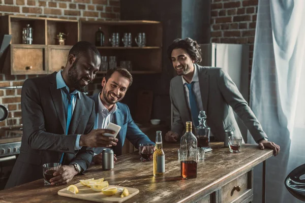 Усміхнені друзі чоловічої статі в костюмах, використовуючи смартфон і п'ючи алкогольні напої під час вечірки вдома — стокове фото