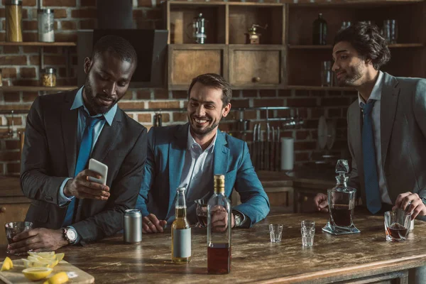 Handsome young men in suits using smartphone and drinking alcohol beverages while partying at home — Stock Photo