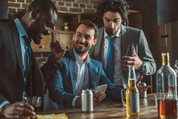 Alegres hombres multiétnicos en trajes usando smartphone y bebiendo bebidas alcohólicas juntos - foto de stock