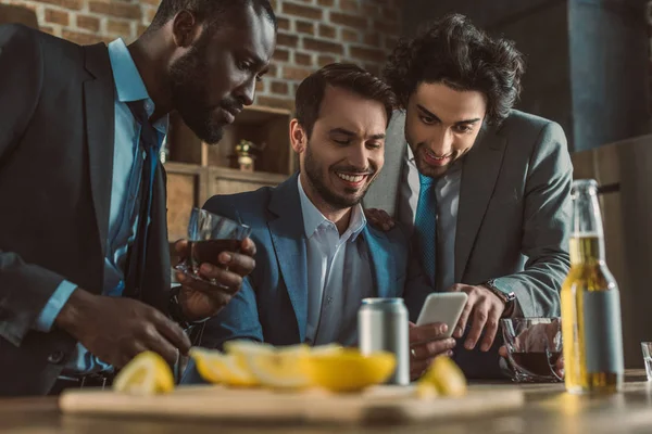 Uomini multietnici sorridenti che usano smartphone e bevono whisky insieme — Foto stock