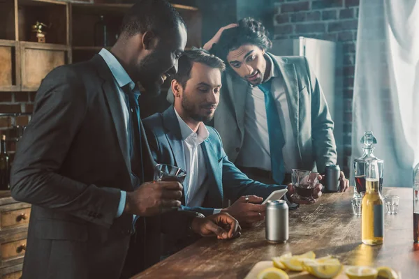 Tres hombres jóvenes en trajes mirando el teléfono inteligente mientras beben whisky juntos - foto de stock